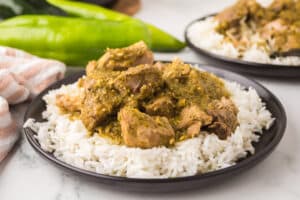 A plate of white rice topped with pieces of pork in a green chile sauce, with green chiles and another plate of the same dish in the background.