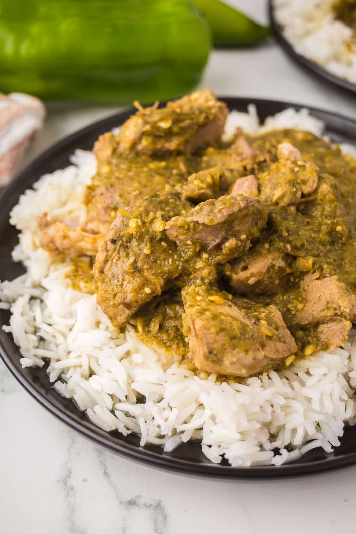 A plate of white rice topped with chunks of meat covered in a green chili sauce. A green chili pepper and another plate of food are in the background.