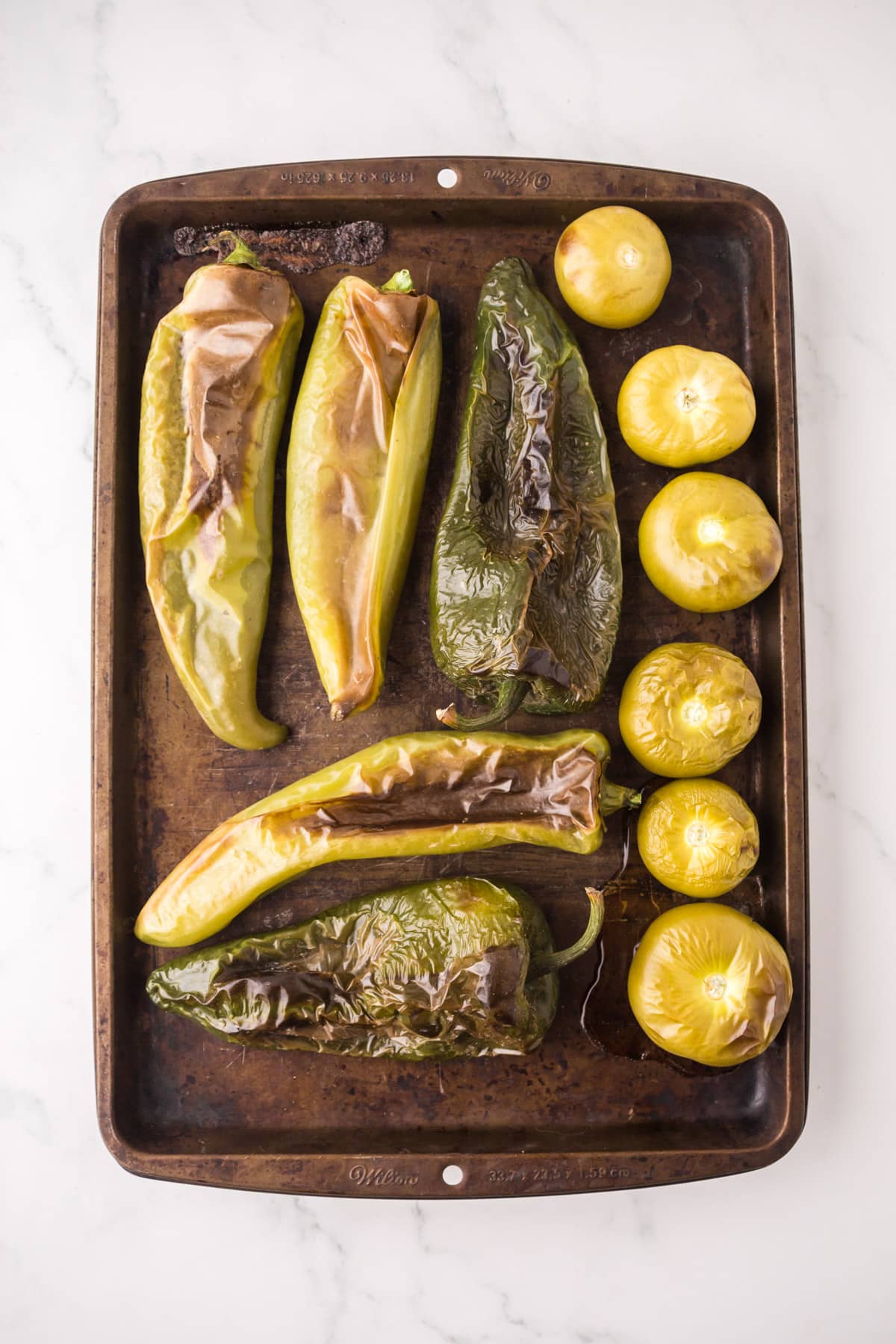 A baking sheet with roasted green peppers and several yellow-green tomatillos, all with blistered skins, on a white marble surface.