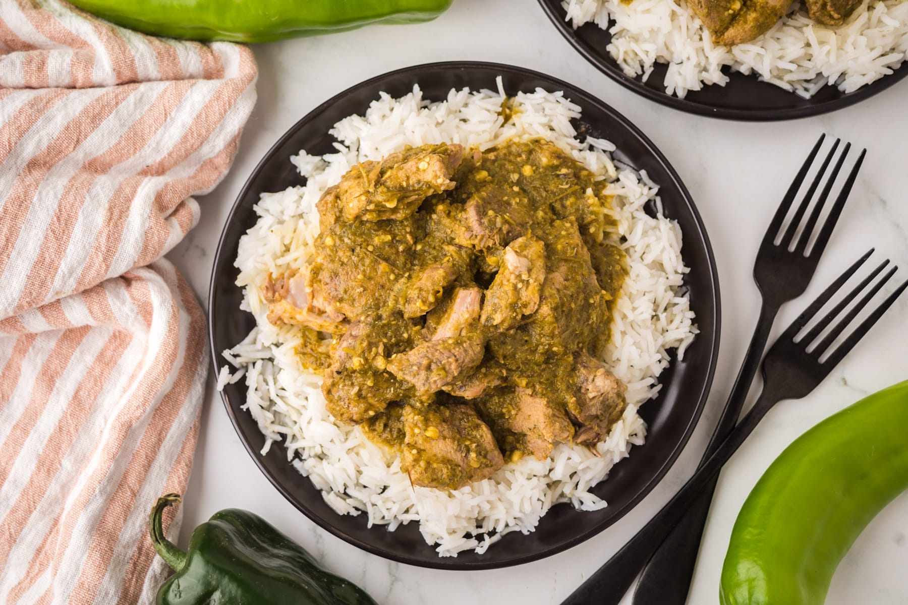 A black plate filled with white rice topped with green chile pork stew. The plate is on a white surface, surrounded by green chilies, a striped napkin, two black forks, and another similar plate partially visible.