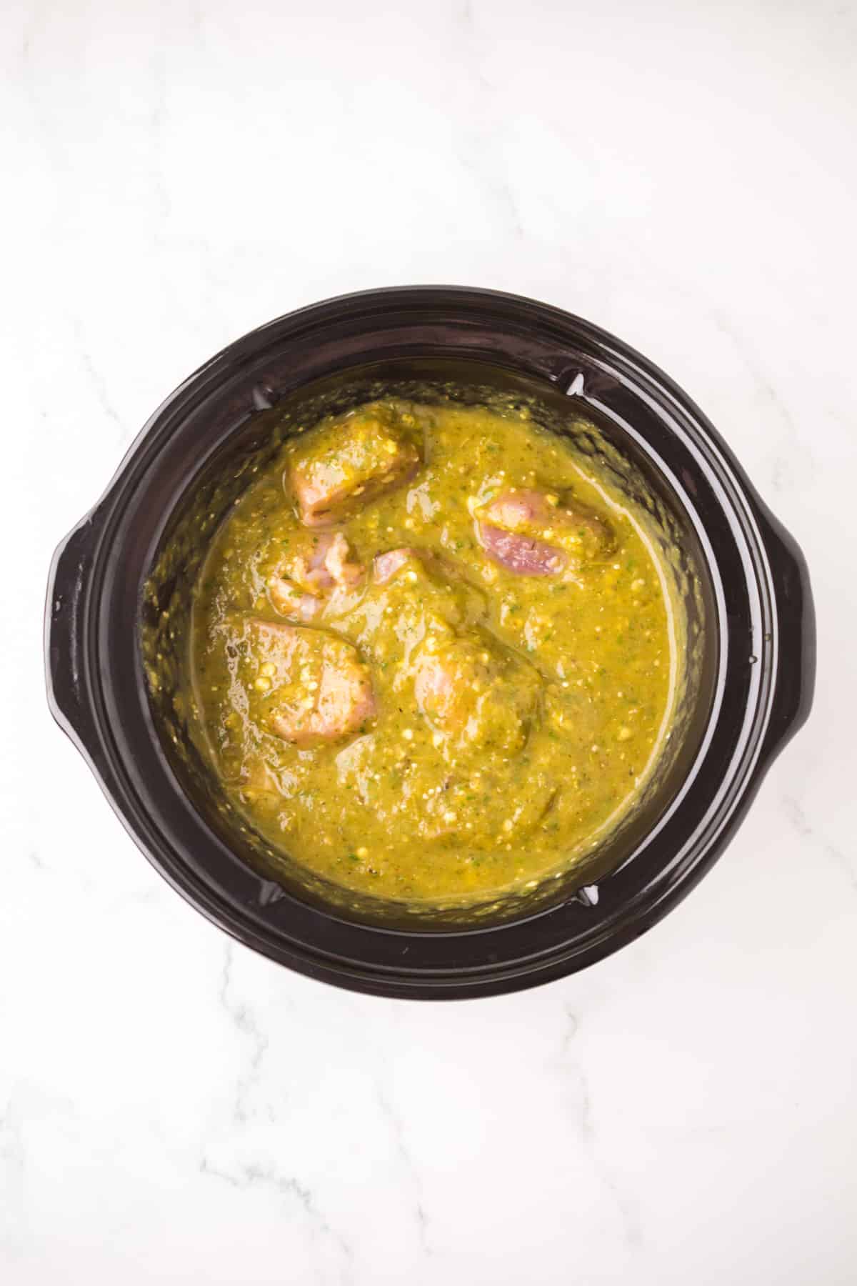 A black slow cooker filled with chunks of raw meat covered in green salsa on a white marble countertop.