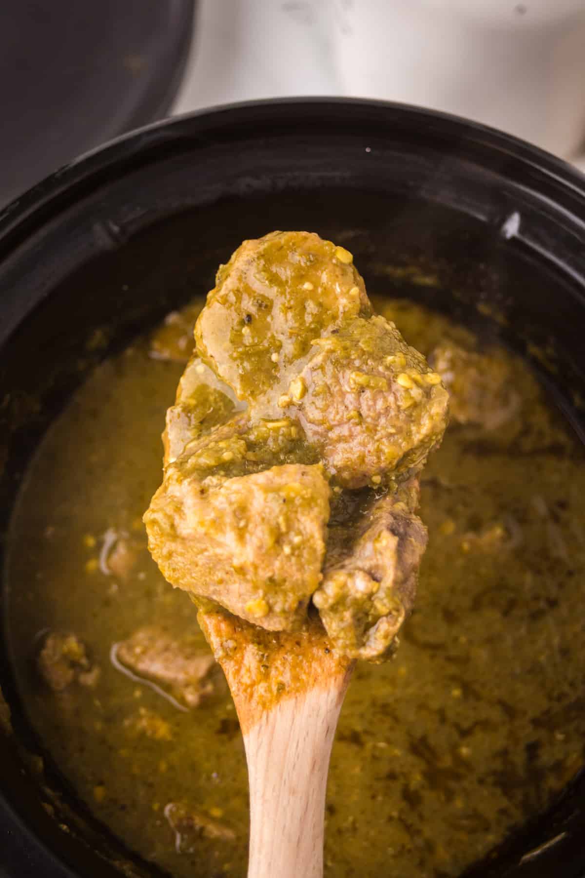 A wooden spoon lifts chunks of beef in a thick, yellow-green curry sauce from a black pot.