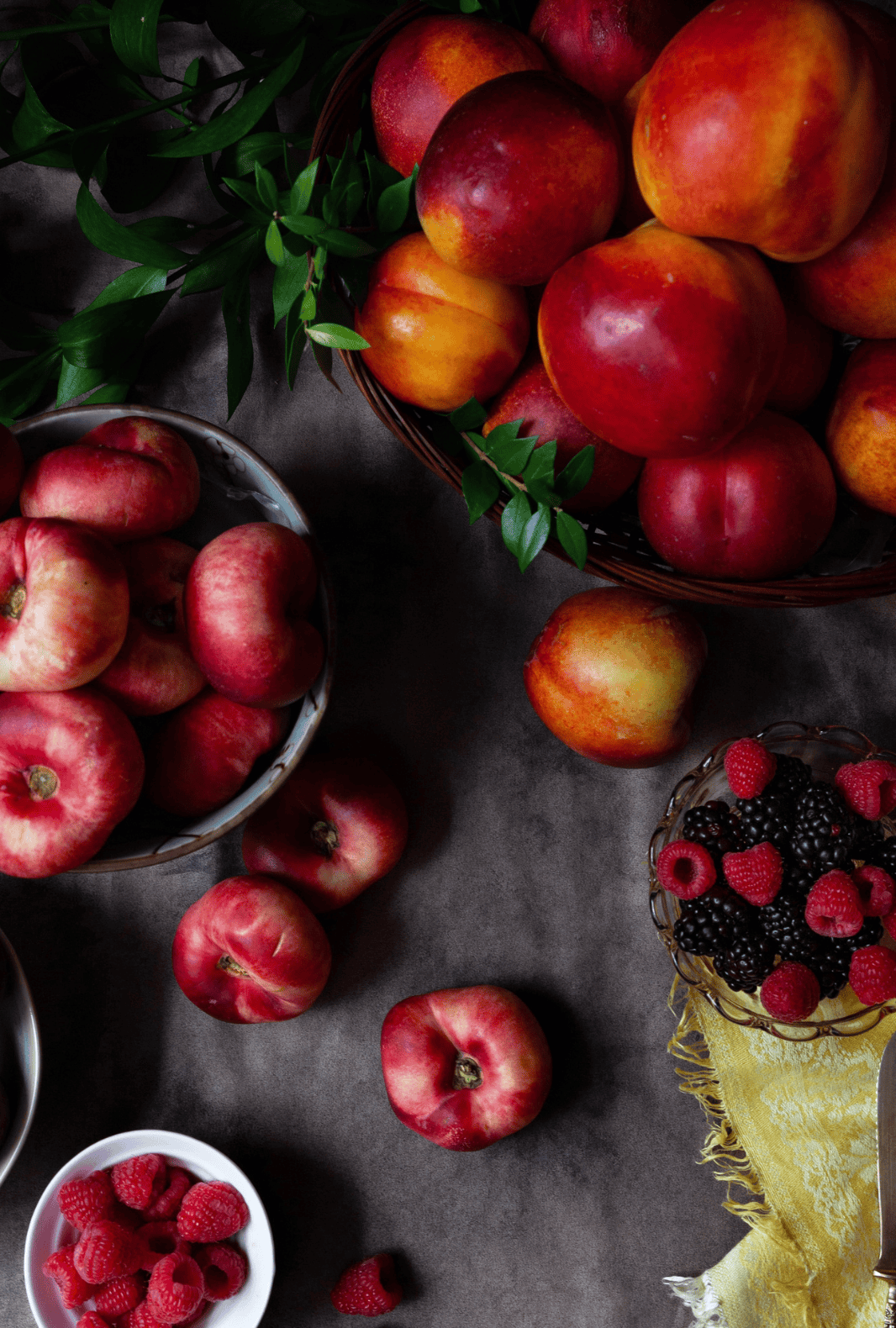 Flat-lay of various fresh fruits including peaches, nectarines, raspberries, and blackberries on a dark surface with a yellow napkin, knife, and blessthismessplease.com label at the bottom.