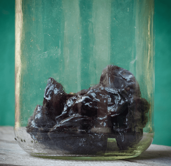 A close-up of a glass jar containing several dark, glossy preserved plums or prunes, placed on a wooden surface with a green background.