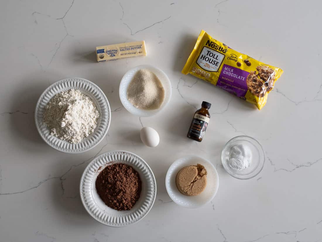Top-down view of baking ingredients on a white counter: flour, cocoa powder, butter, sugar, brown sugar, an egg, vanilla extract, baking soda in a small bowl, and a bag of Nestlé Toll House milk chocolate chips.