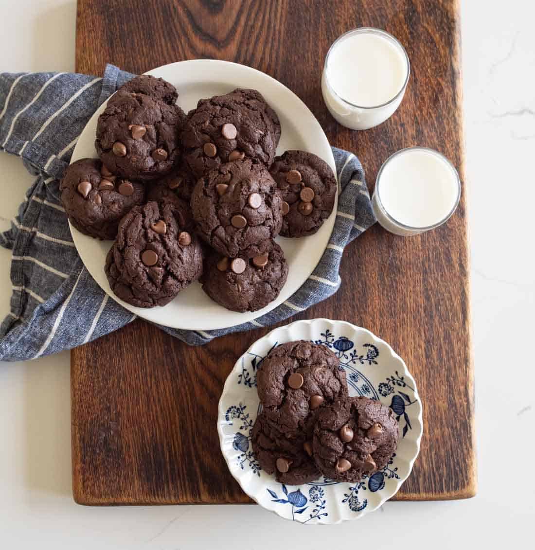 A plate of chocolate cookies with chocolate chips sits on a wooden board next to two glasses of milk. A small floral plate holds two more cookies, and a striped cloth is partially under the main plate.