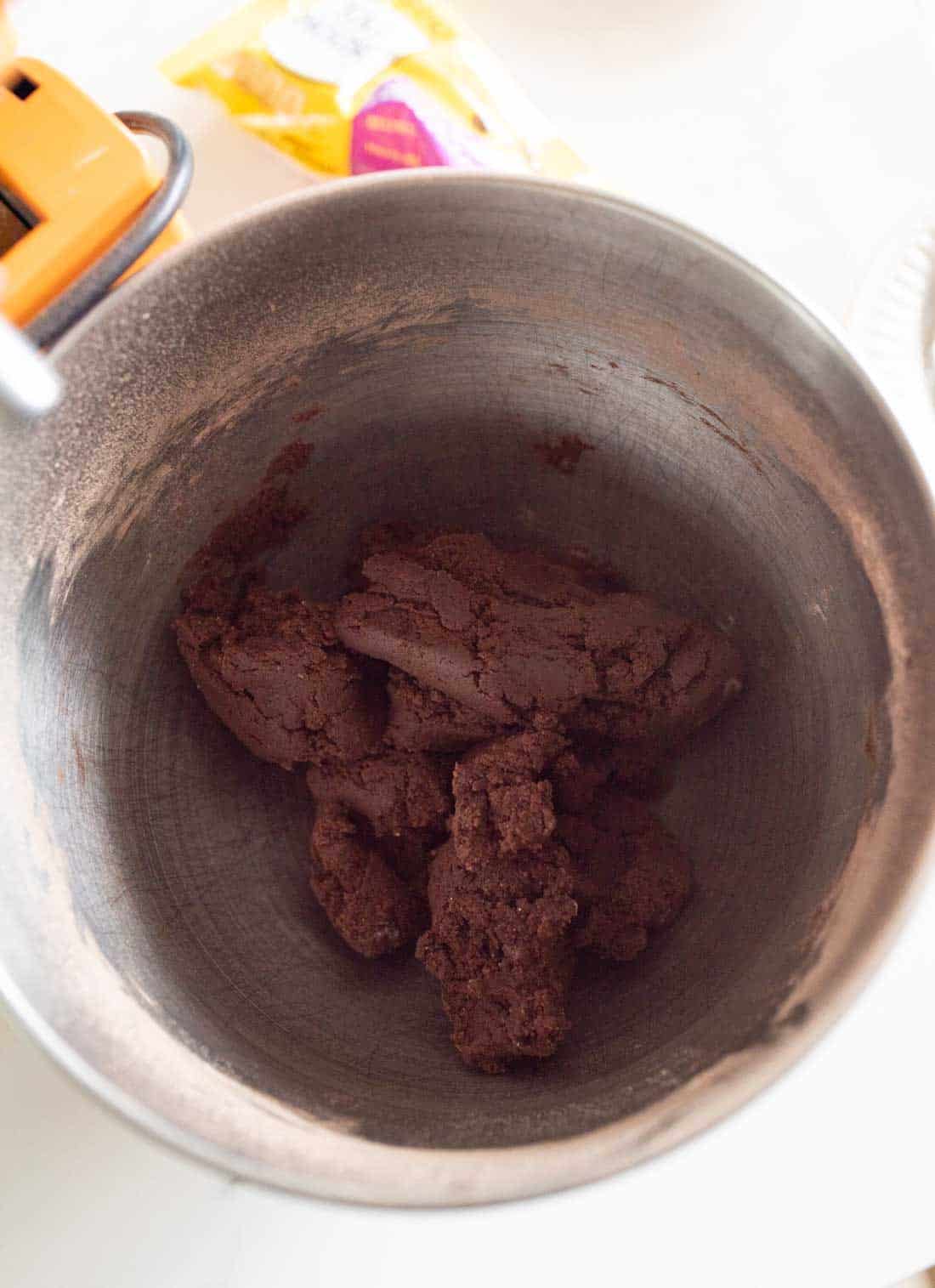 A metal mixing bowl containing dark chocolate cookie dough, with a stand mixer attachment partially visible on the side and a yellow package in the background.