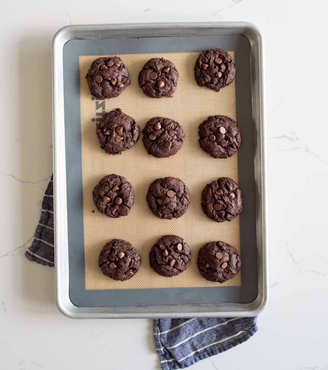 A baking sheet with 12 chocolate cookies topped with chocolate chips, arranged in rows on a silicone baking mat. The tray is on a white surface with a striped kitchen towel partially visible underneath.