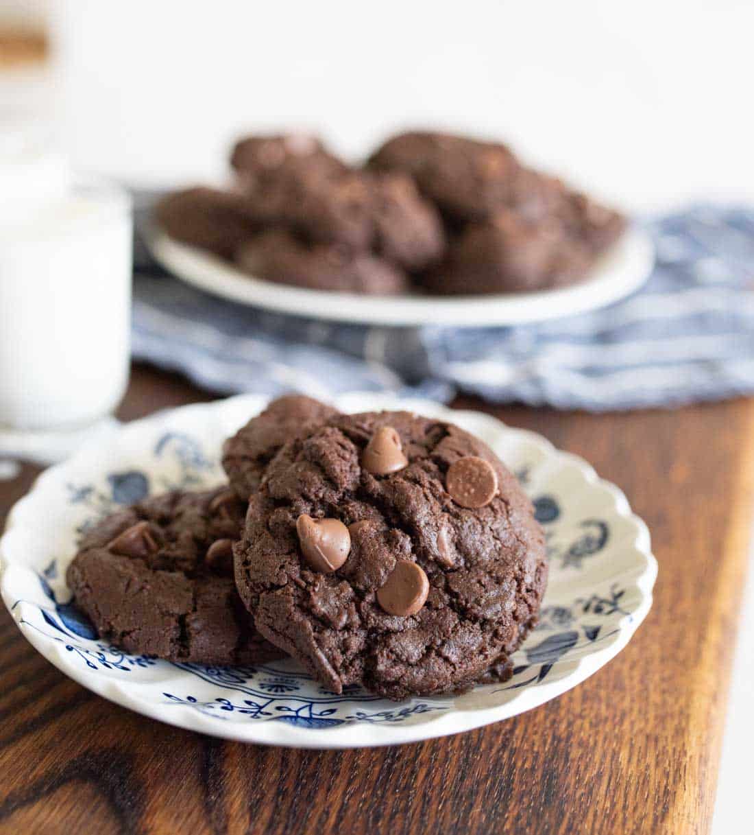 A plate of double chocolate chip cookies sits on a wooden table, with a glass of milk and more cookies on a plate in the background. The cookies are rich, dark, and studded with chocolate chips.