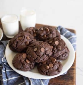 A plate of chocolate chocolate-chip cookies sits on a blue plaid cloth, with two glasses of milk in the background. The cookies are thick and soft with visible chocolate chips.