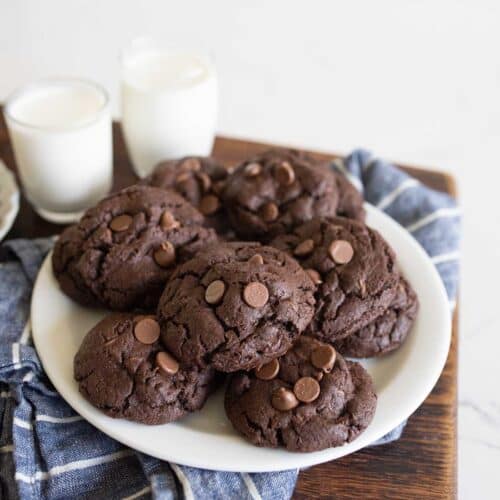 A plate of chocolate chocolate-chip cookies sits on a blue plaid cloth, with two glasses of milk in the background. The cookies are thick and soft with visible chocolate chips.