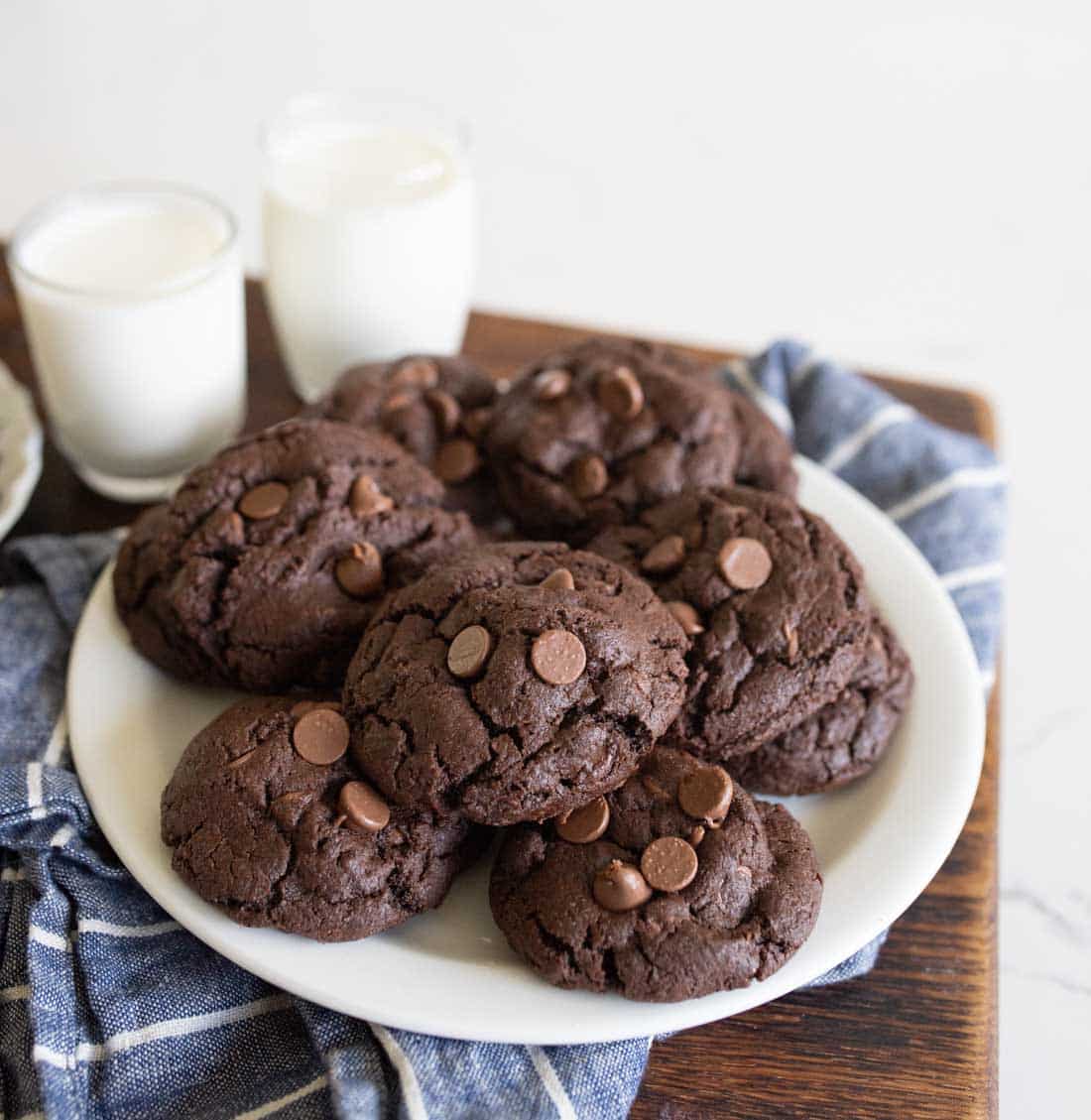 A plate of chocolate chocolate-chip cookies sits on a blue plaid cloth, with two glasses of milk in the background. The cookies are thick and soft with visible chocolate chips.