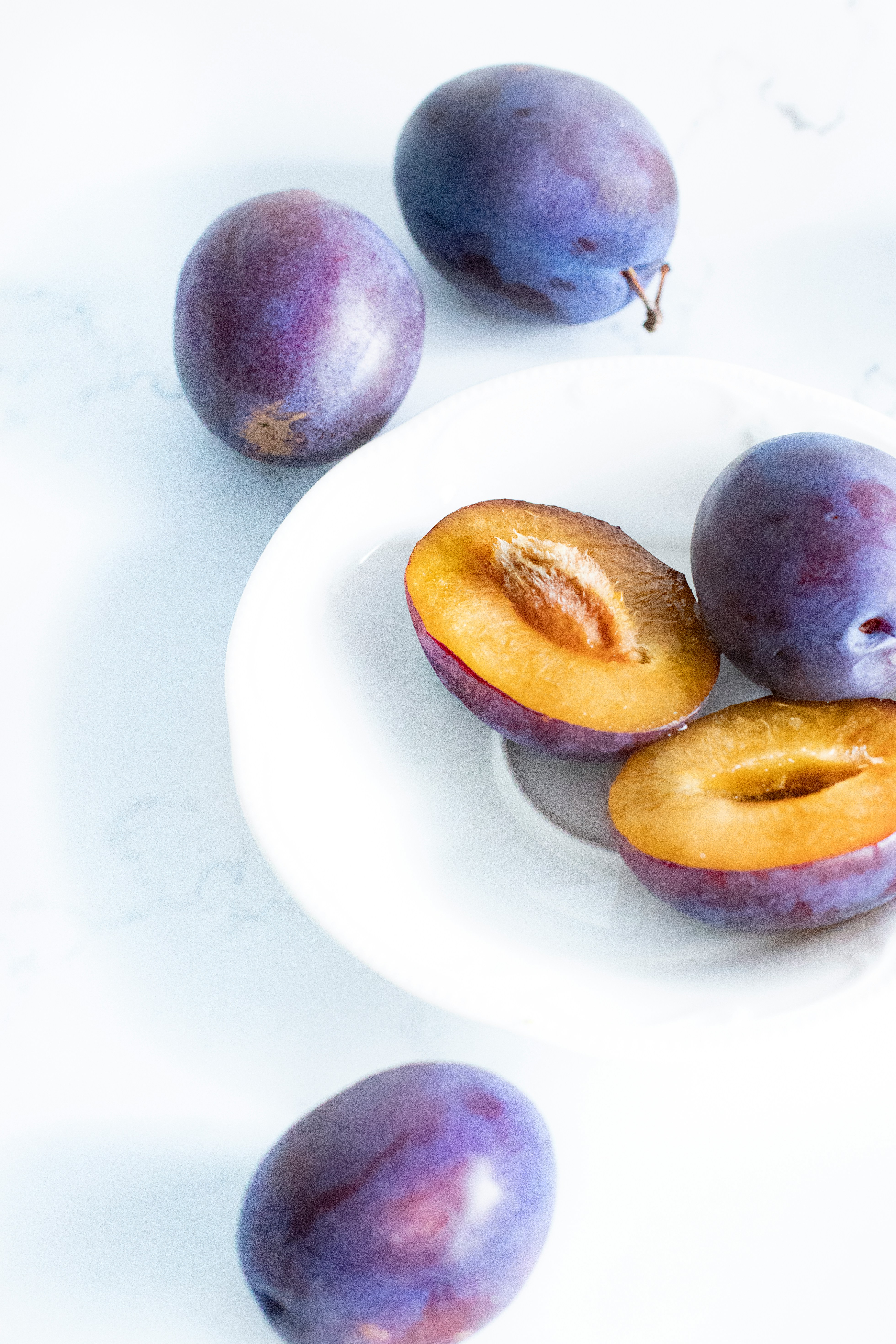 Three whole purple plums and one plum cut in half with the pit exposed are arranged on a white plate, with another whole plum placed nearby on a white, bright background.