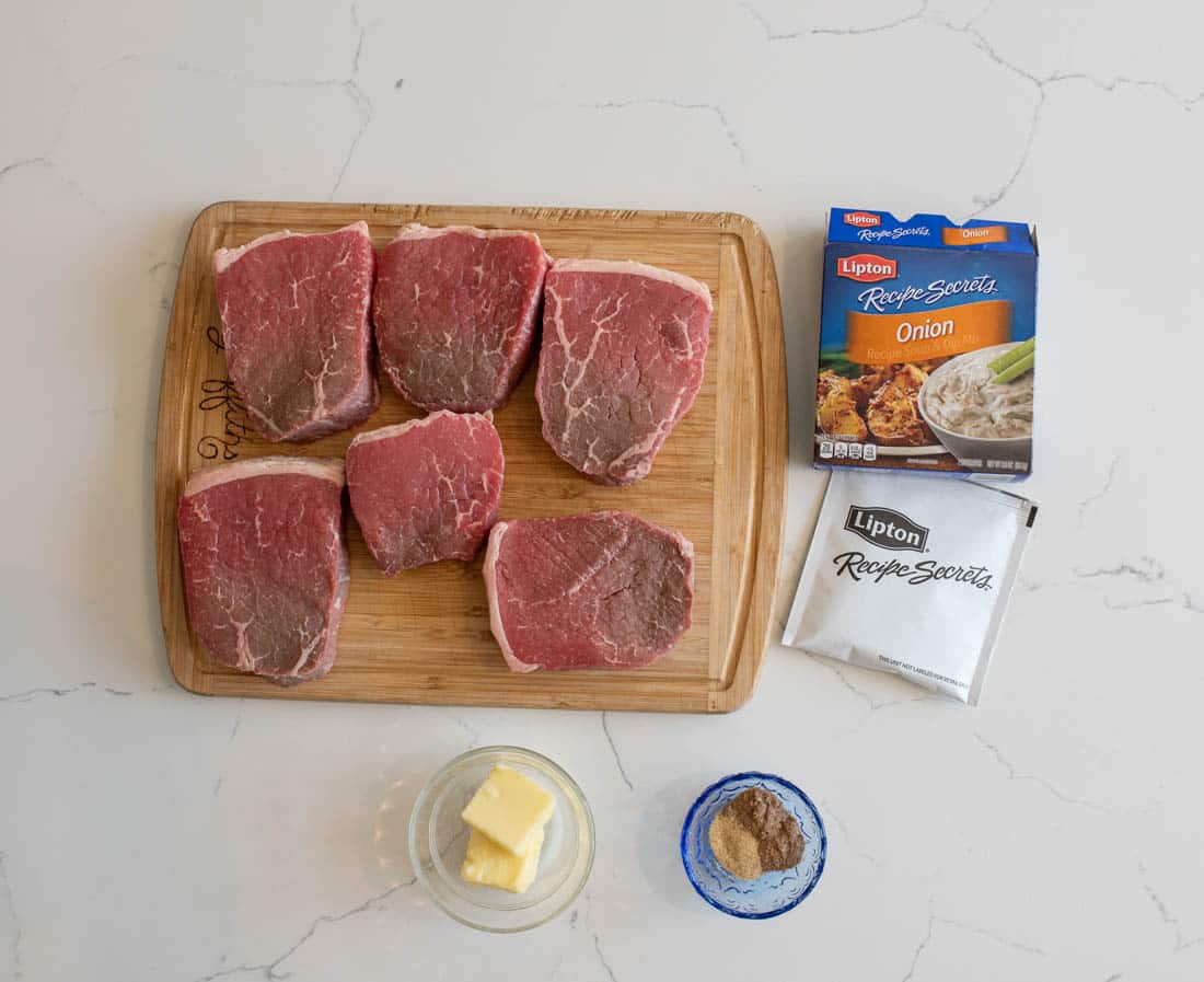 Six raw beef steaks on a wooden cutting board, with a packet and box of Lipton Recipe Secrets onion soup mix, a small bowl of butter, and a small bowl of seasoning, all on a white surface.