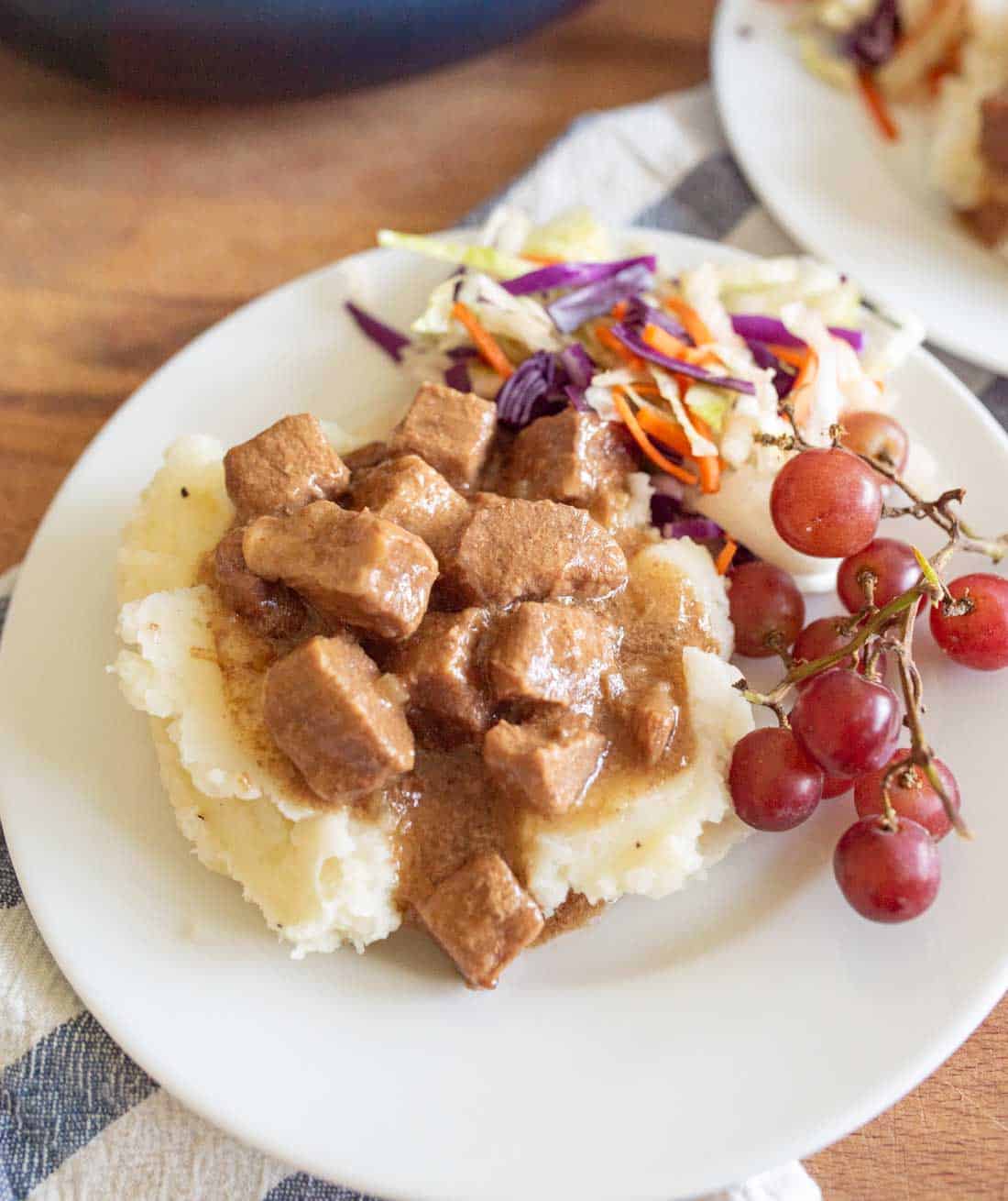 A plate of mashed potatoes topped with beef stew, served with a side of coleslaw and a small bunch of red grapes.