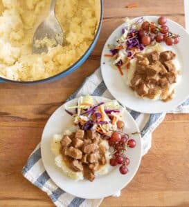 Two plates with mashed potatoes topped with brown stew, colorful coleslaw, and red grapes sit on a cloth. A pot of mashed potatoes with a serving spoon is nearby on a wooden surface.