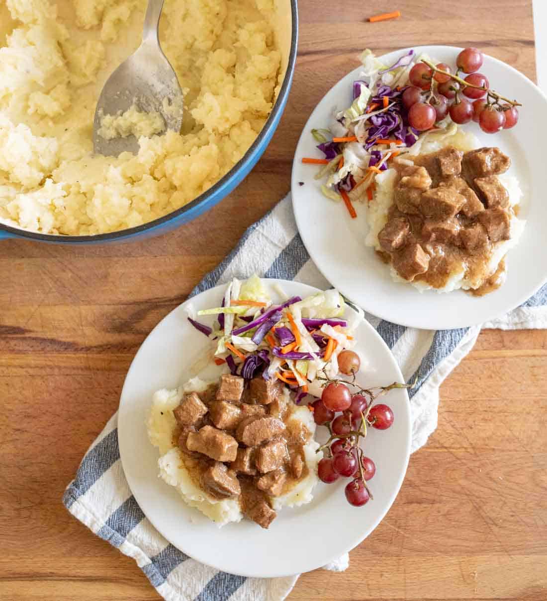 Two plates with mashed potatoes topped with brown stew, colorful coleslaw, and red grapes sit on a cloth. A pot of mashed potatoes with a serving spoon is nearby on a wooden surface.