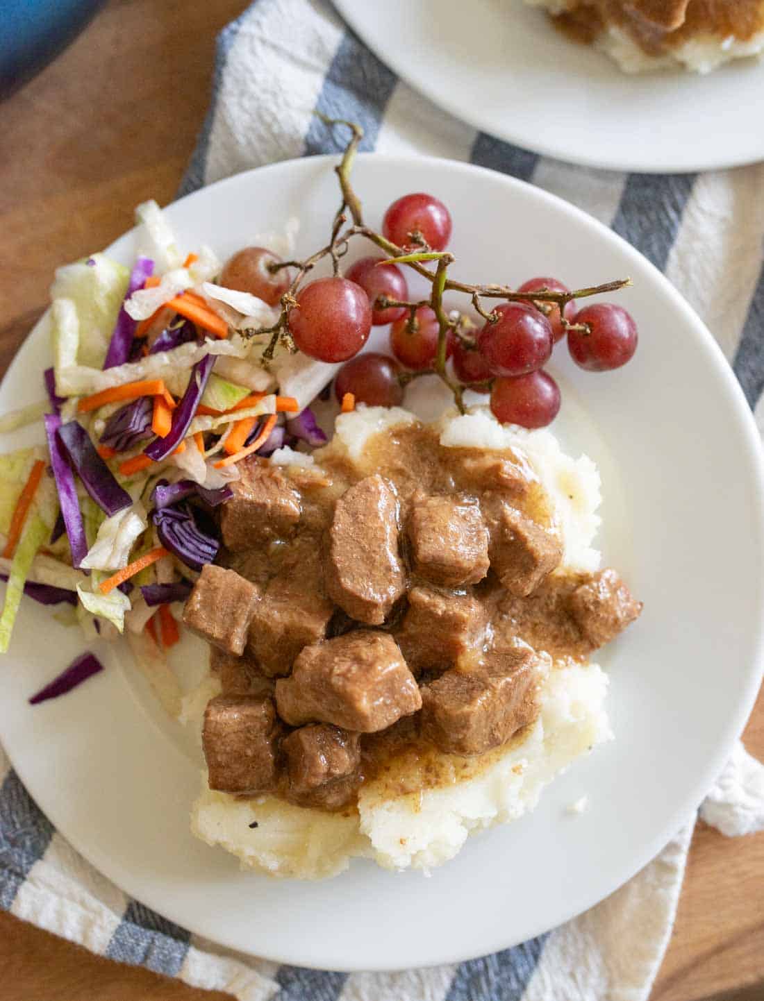 A white plate with mashed potatoes topped with beef stew, a side of coleslaw, and a small bunch of red grapes, set on a striped cloth.