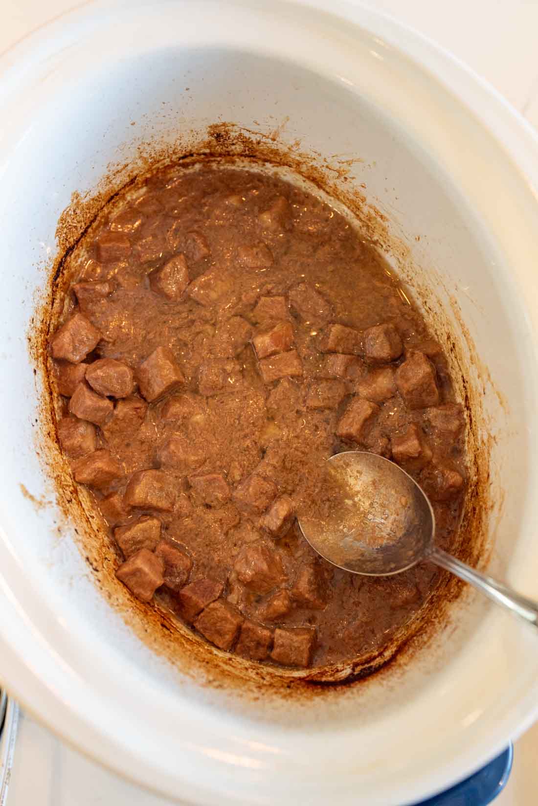 Cubed beef simmering in a brown gravy inside a white slow cooker, with a metal spoon resting on the side.